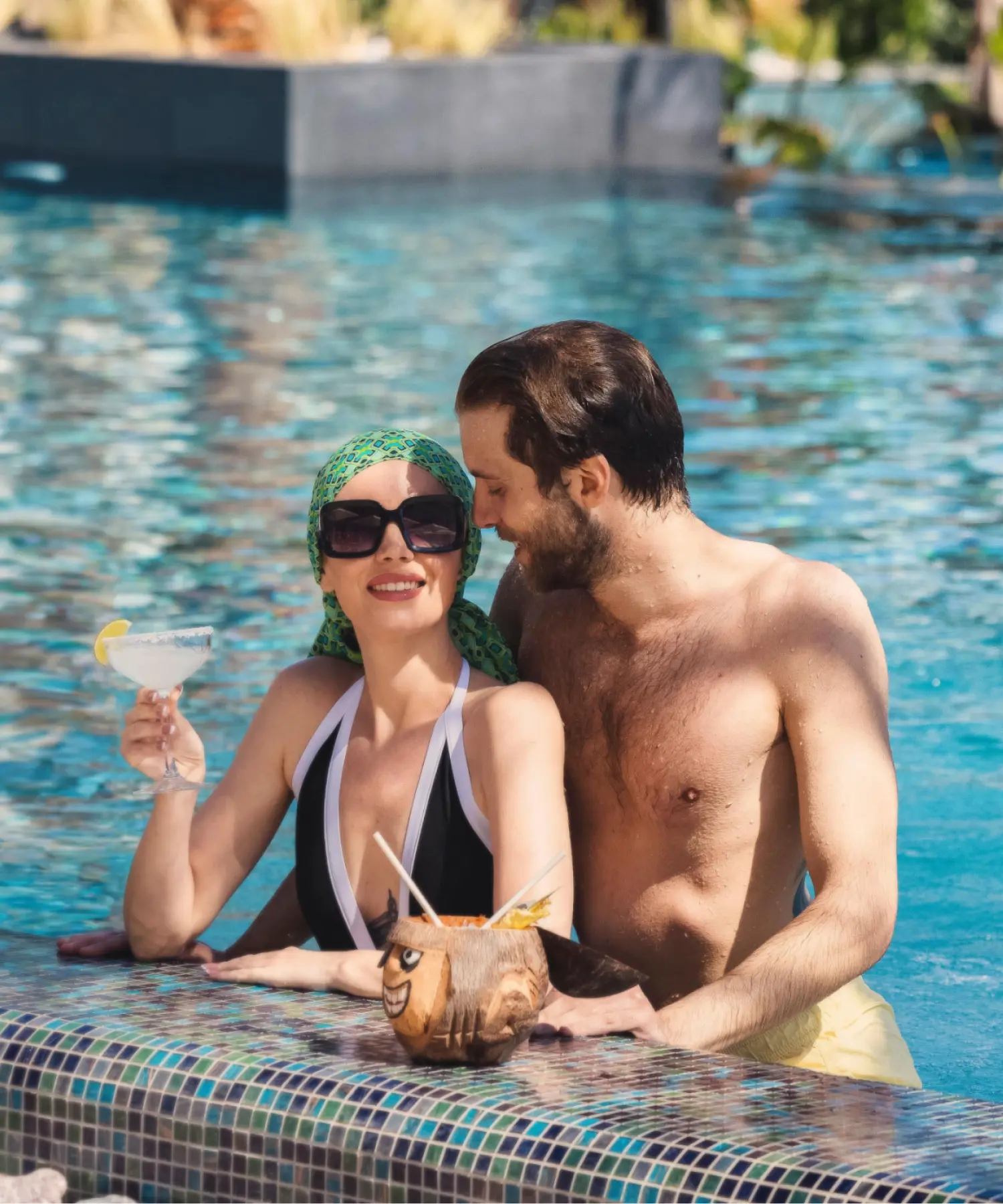 A couple relaxing together in a leisure swimming pool, with the woman holding a cocktail glass and wearing sunglasses and a headscarf while resting against the pool edge.